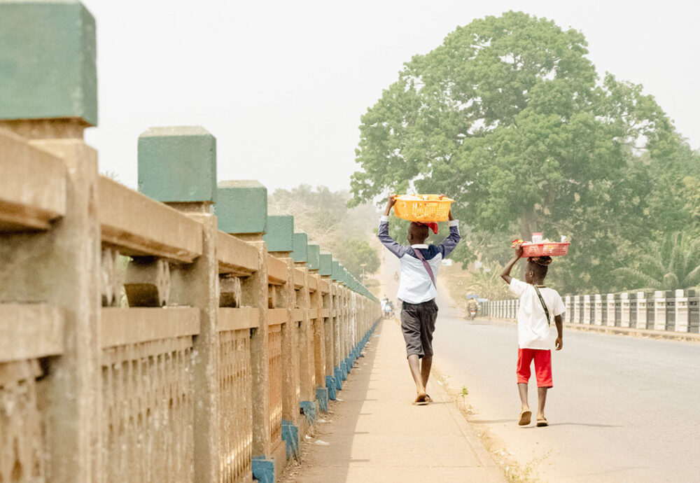 Two boys cross a bridge in Kambia, Sierra Leone, carrying an array of items for sale. In the country’s Eastern Province, where Baindu lives, 33% of children under 18 have experienced some form of child trafficking.